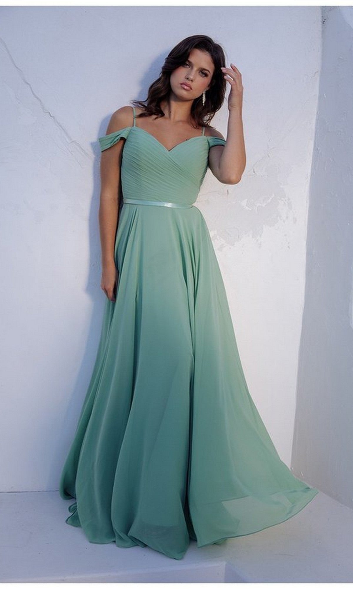 A woman in the sage Eureka 7611 long prom dress, featuring an off-the-shoulder chiffon design and ruched fitted bodice, stands against a white textured wall.